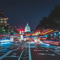 A nighttime city scene with light trails from traffic leading toward a well-lit domed building, likely representing Washington D.C.''s Capitol.