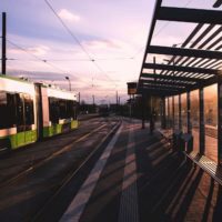 A tram station at sunset with an arriving tram, empty platform, shelter, and tracks. Shadows cast by the setting sun create a serene atmosphere.