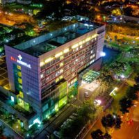 Aerial view of a hospital building illuminated at night, surrounded by trees and city lights.