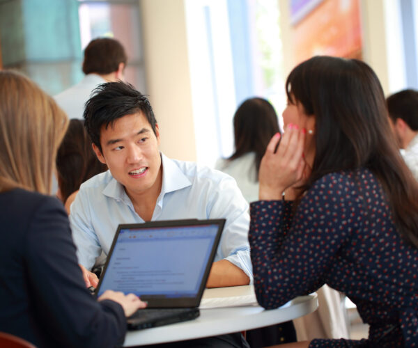 Students talking indoors around a round table with laptops open