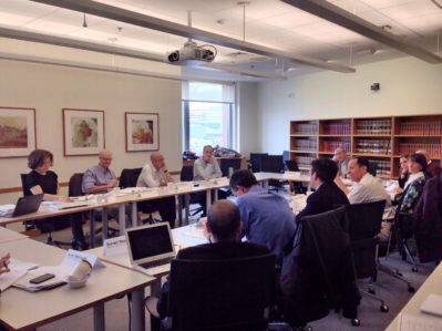 A group of people engaged in a meeting around a large conference table in a well-lit office room with bookshelves and framed art on the walls.