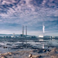 Industrial landscape with smokestacks and a cloudy sky, reflecting on a wet, possibly polluted surface, suggesting themes of industrialization or environmental impact.