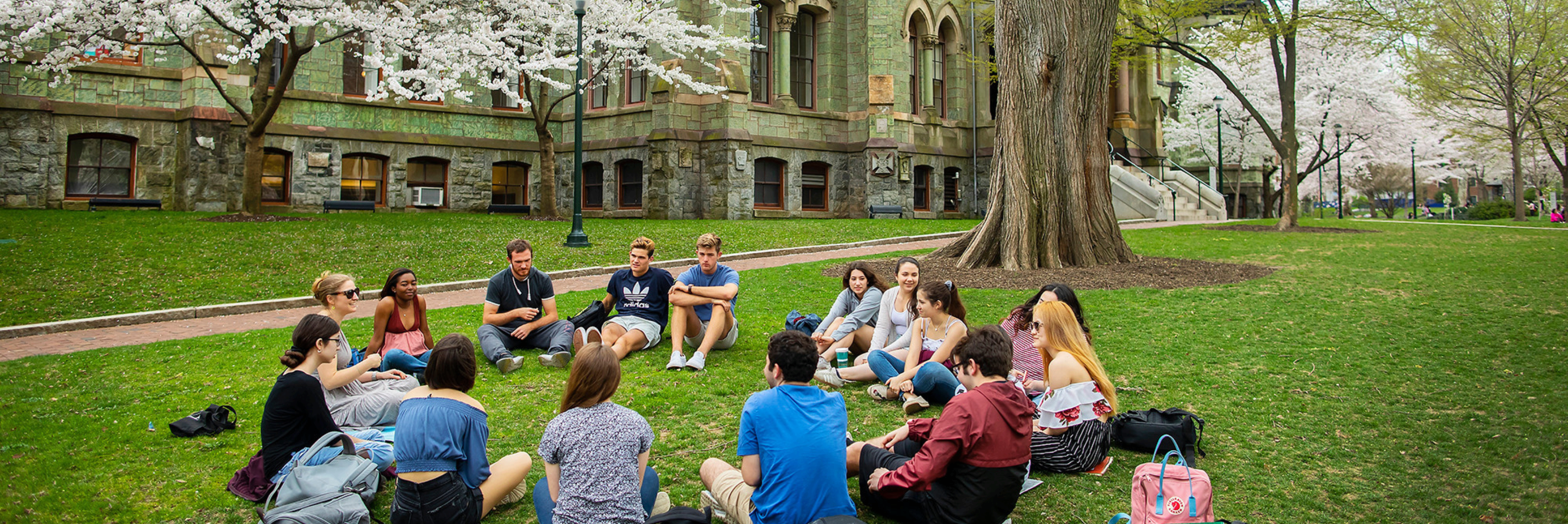 Student Club Engagement Students gathered in a circle chatting on the grass on campus during the spring time with cherry blossoms blooming in the background