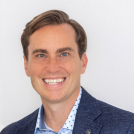 Tyler Wry smiling in a white button down shirt with a blue pattern and a navy suit jacket against a white wall