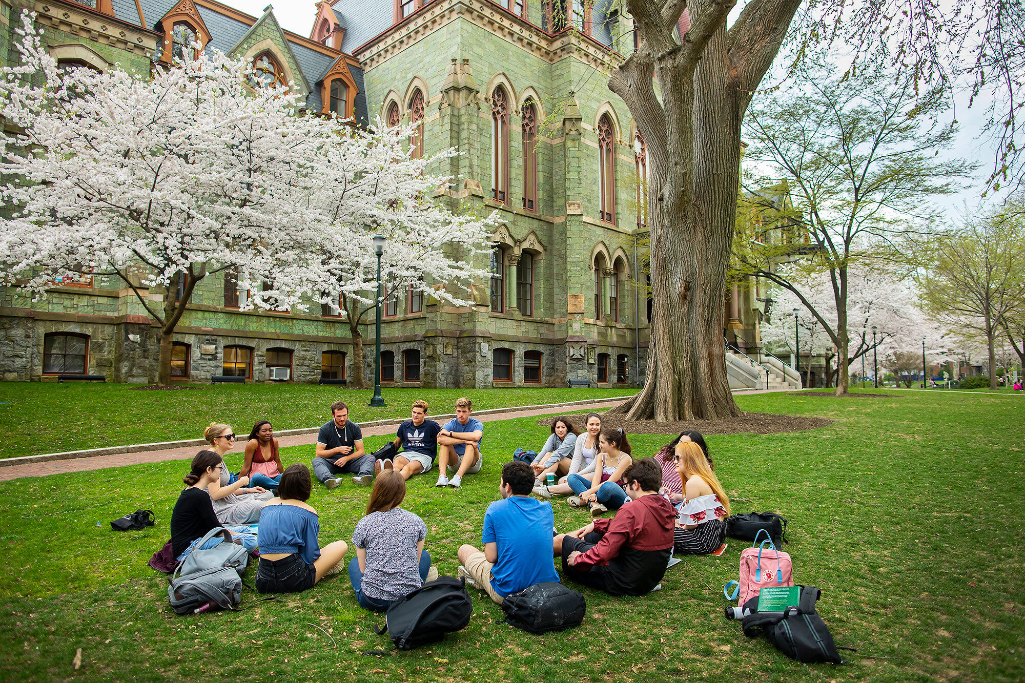College Hall University Of Pennsylvania Students gathered in a circle chatting on the grass on campus during the spring time with cherry blossoms blooming in the background