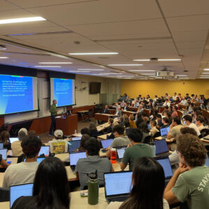 2023 Climate Prof Img 1 Image of a packed tiered classroom of students, some with laptops open, watching a speaker present research from the front of the classroom.