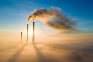 Aerial,View,Of,Coal,Power,Plant,High,Pipes,With,Black Aerial view of three coal power plant pipes with black smoke emerging from two and blooming backwards into the blue sky. Below are clouds from above, with the shadow of the smoke stacks.
