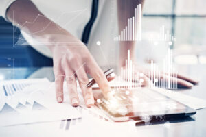 Picture,Female,Hand,Touching,Modern,Tablet.investment,Manager,Working,New,Private A hand holding a pen hovers above a tablet set on a table surrounded by papers with graphs on them. Person in background wears a white shirt and blue tie and blue pants, standing in front of a window. Graphs are overlaid over the image.