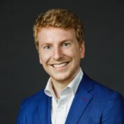 Dan G Smiling person with short blonde hair wearing a blue suit and white shirt, against a dark background. Headshot.