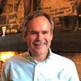 Frank D Headshot of a smiling person with a mustache, wearing a light-colored shirt, standing in front of a rustic brick and wood background.