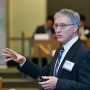 JPM A person wearing glasses and a suit is speaking or presenting in a formal setting, possibly a conference or lecture.