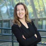 Sandra S Sandra Schafhäutle, a white woman with blonde hair wearing a black blazer and shirt, smiles into the camera with her arms crossed in front of a railing and a glass building's facade.