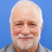 Steven K Headshot of a person with short white hair and a beard, smiling against a blue background.