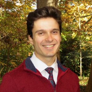 A person smiling in a forest setting, wearing a red sweater over a white shirt and patterned tie, amidst autumn foliage.