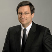 William Laufer Headshot Professional headshot of William Laufer in a white button down, dark tie, and black suit jacket looking off to the side in front of a grey backdrop.