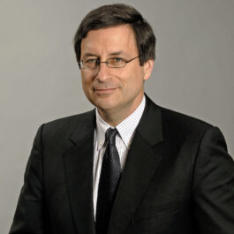 Professional headshot of William Laufer in a white button down, dark tie, and black suit jacket looking off to the side in front of a grey backdrop.