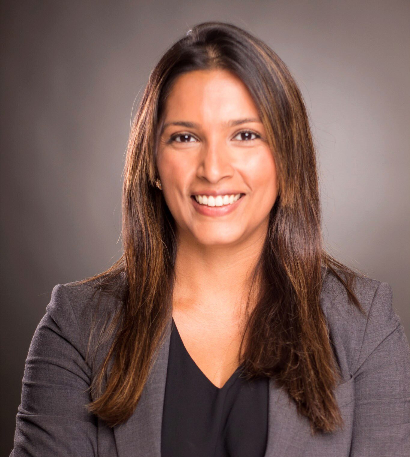 Aarthi Sowrirajan poses for a professional headhot, smiling into the camera in a black blouse and grey blazer in front of a grey backdrop.