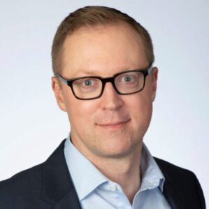 1570049150498 Headshot of a person with short light hair, wearing glasses, a light blue shirt, and a dark blazer, set against a plain background.