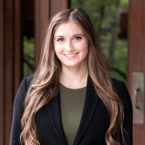 Jess Stokes smiling in a black blazer and an olive green top outside with trees visible through the reflection on a door.