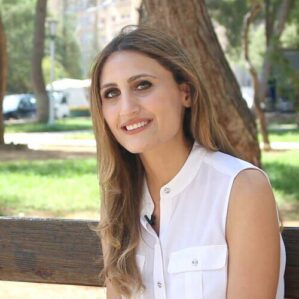N.Ismail+Photo+1 A person with long hair wearing a sleeveless white shirt is sitting on a bench in a park setting, smiling. Trees and greenery are visible in the background.