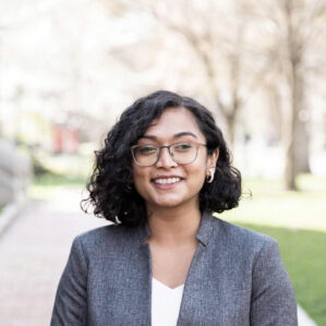 Rishika Ghosh smiling in a grey blazer and white top outside on a sunny day with trees in the background.