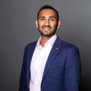 Shrinidhi Rao smiling in a navy blue suit and a white button down, posed in front of a dark grey backdrop.