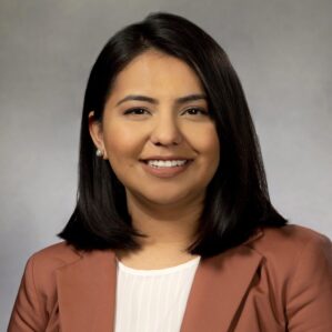 Cynthia Jasso Headshot of a smiling person with shoulder-length dark hair, wearing a brown jacket and white blouse, against a neutral background.
