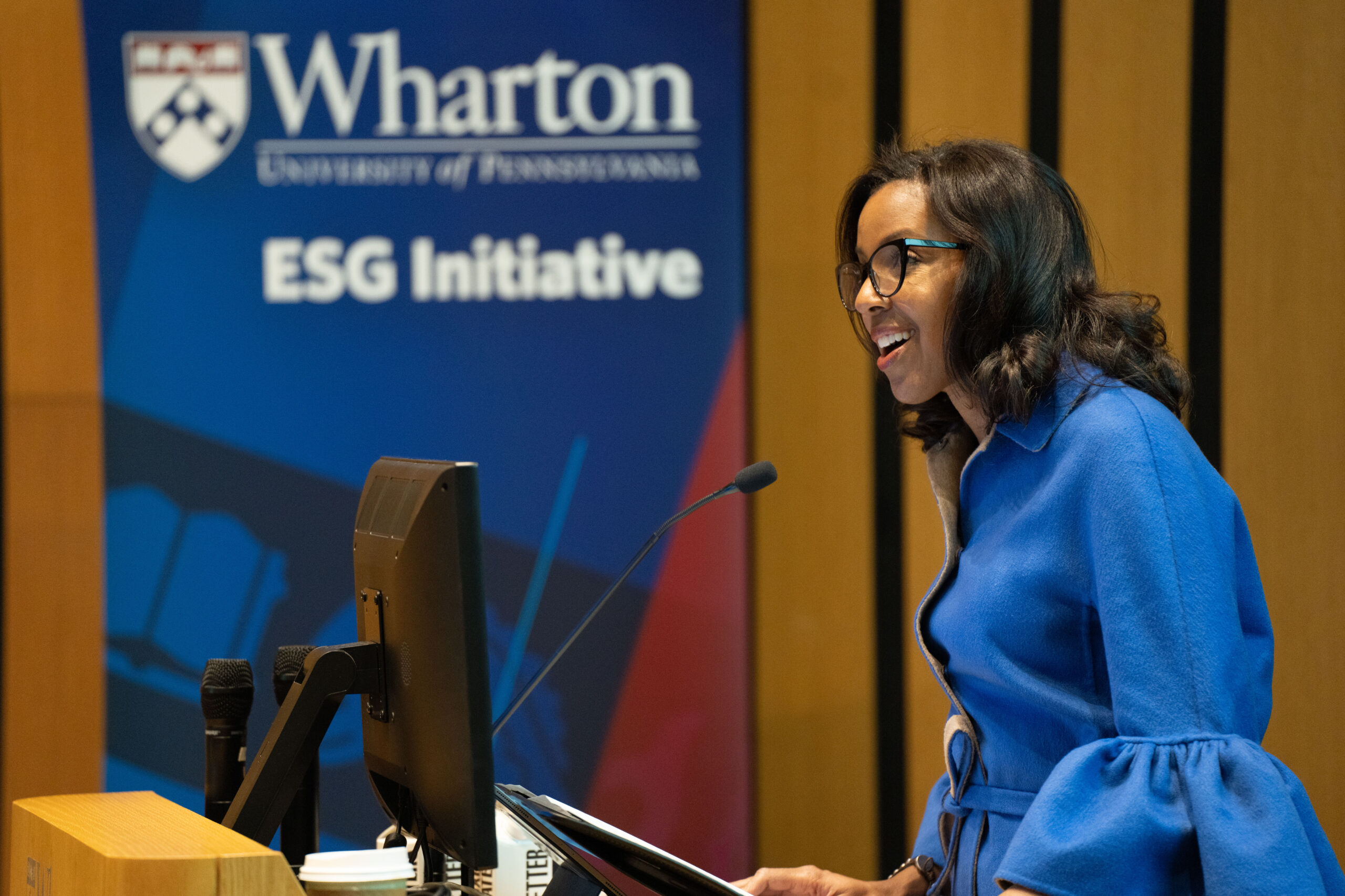 Dean James TMIINT 2024 Dean James in a blue blouse smiling and speaking at a podium with a banner in the background that says "Wharton ESG Initiative"