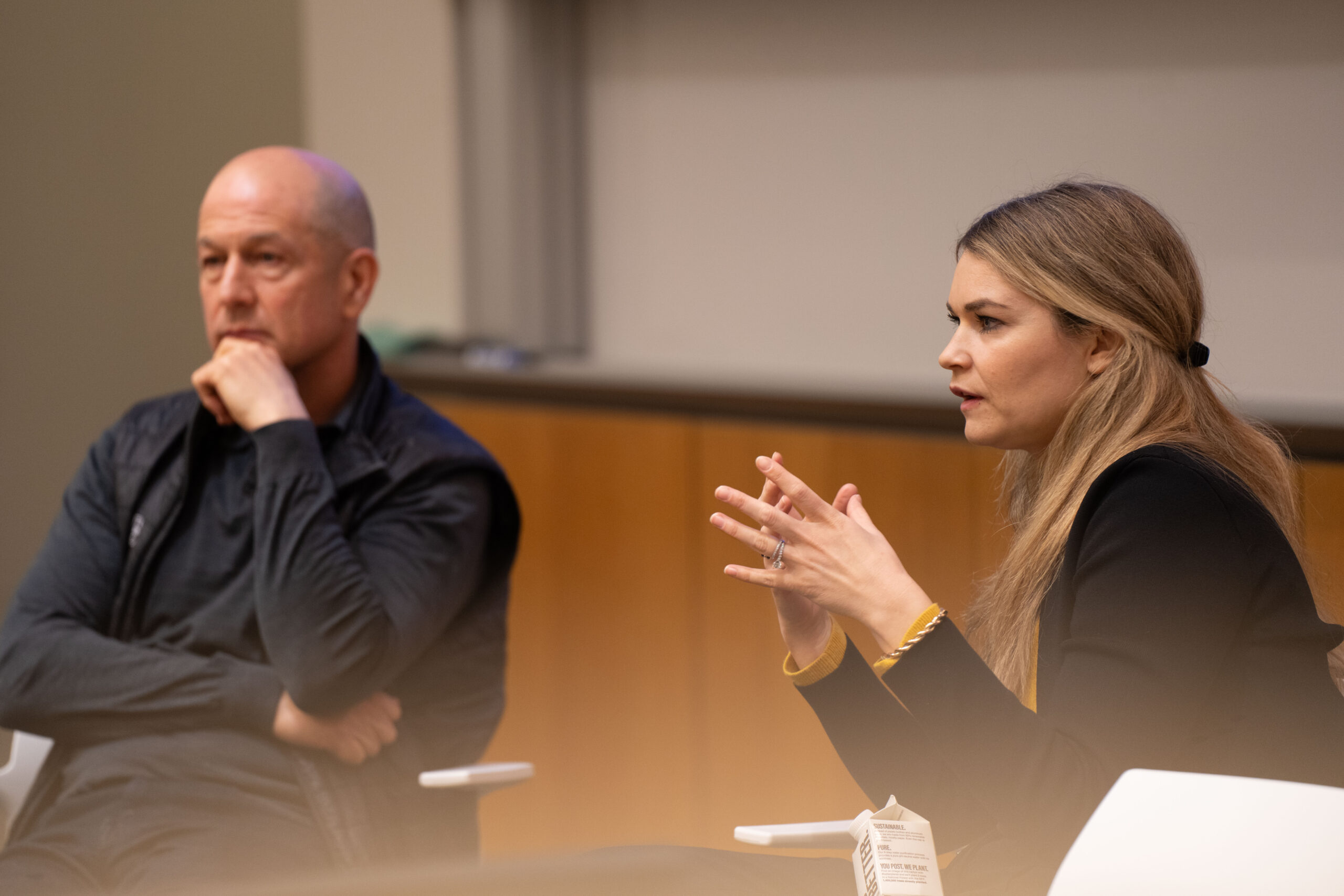 Sarah Norman is seated speaking to an audience out of the shot. Bobby Turner is in the background listening.