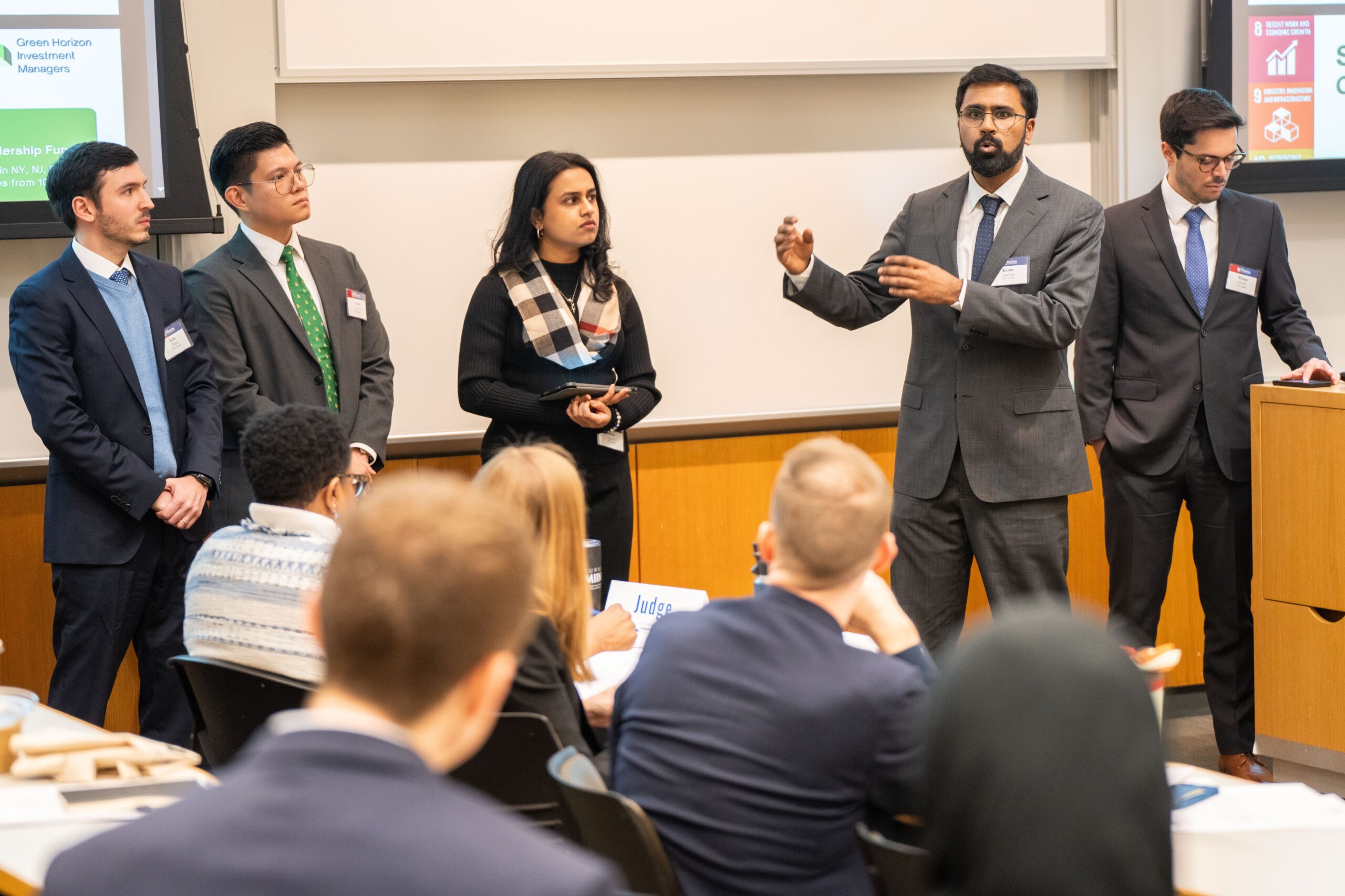 A student team stands at the front of a classroom presenting. You can see the audience and judges in the foreground.