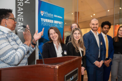 A judge announced the winning team of the 2024 TIPC challenge at a podium and is clapping. The winning team is smiling to the side