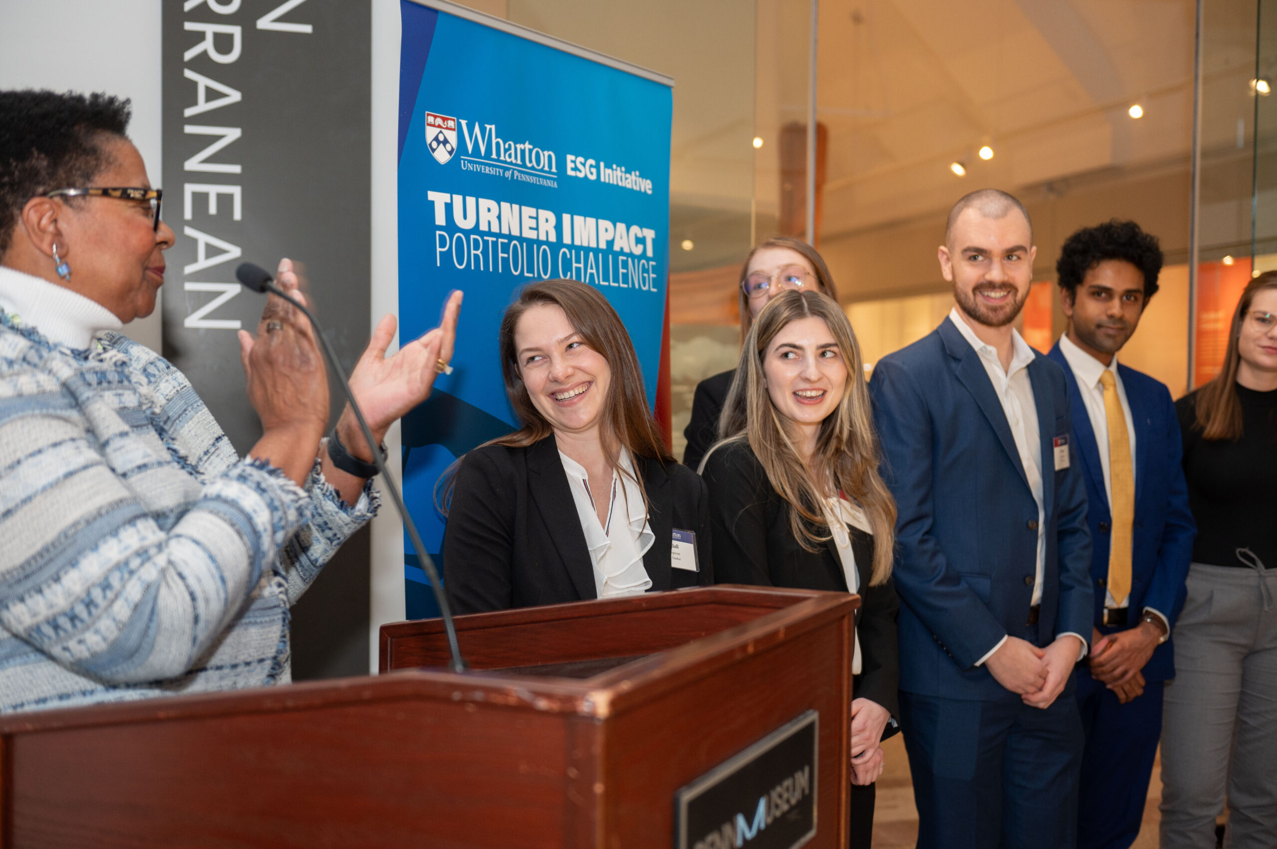 A judge announced the winning team of the 2024 TIPC challenge at a podium and is clapping. The winning team is smiling to the side