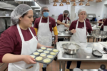 Four chefs stand in a kitchen wearing aprons and red shirts, one holding a tray of cupcakes.