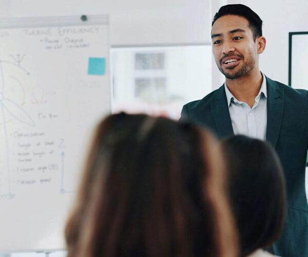 Participant Ntzr A man in a suit presents to a classroom with a large pad with writing in the the background.