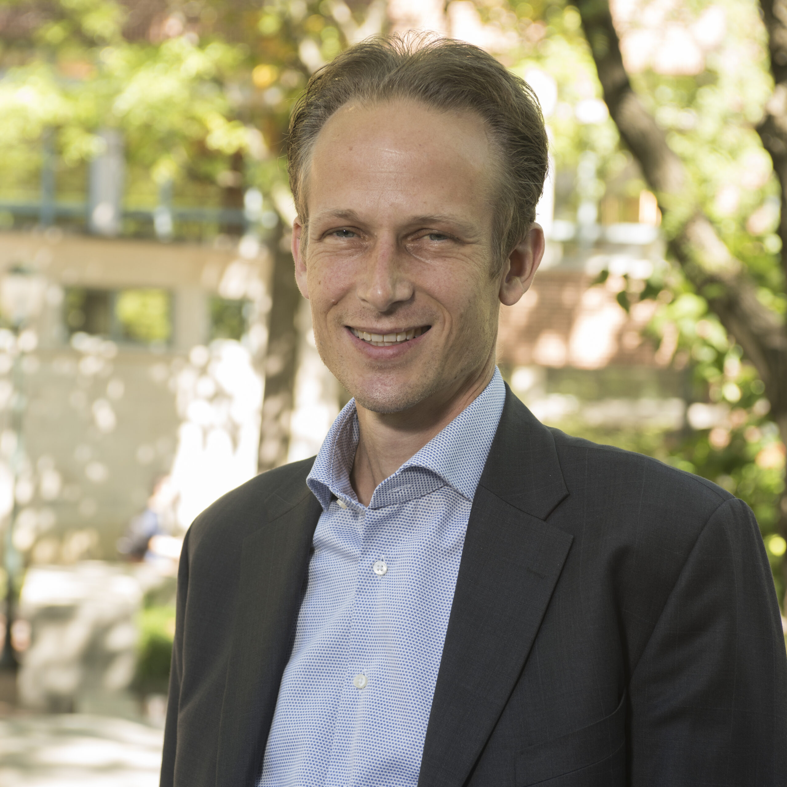 A person wearing a suit standing outdoors, smiling, with sunlight filtering through trees in the background.