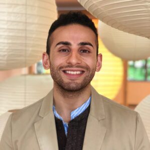 Youssef Kalad smiles for a professional headshot in tan suit jacket with paper lanterns in the background.