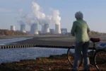 An older woman with a bicycle stares out at a coal fired power plant emitting smoke.