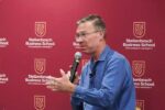 Vit Henisz speaks holding a microphone and gesturing in front of a Stellenboch Business School step-and-repeat.