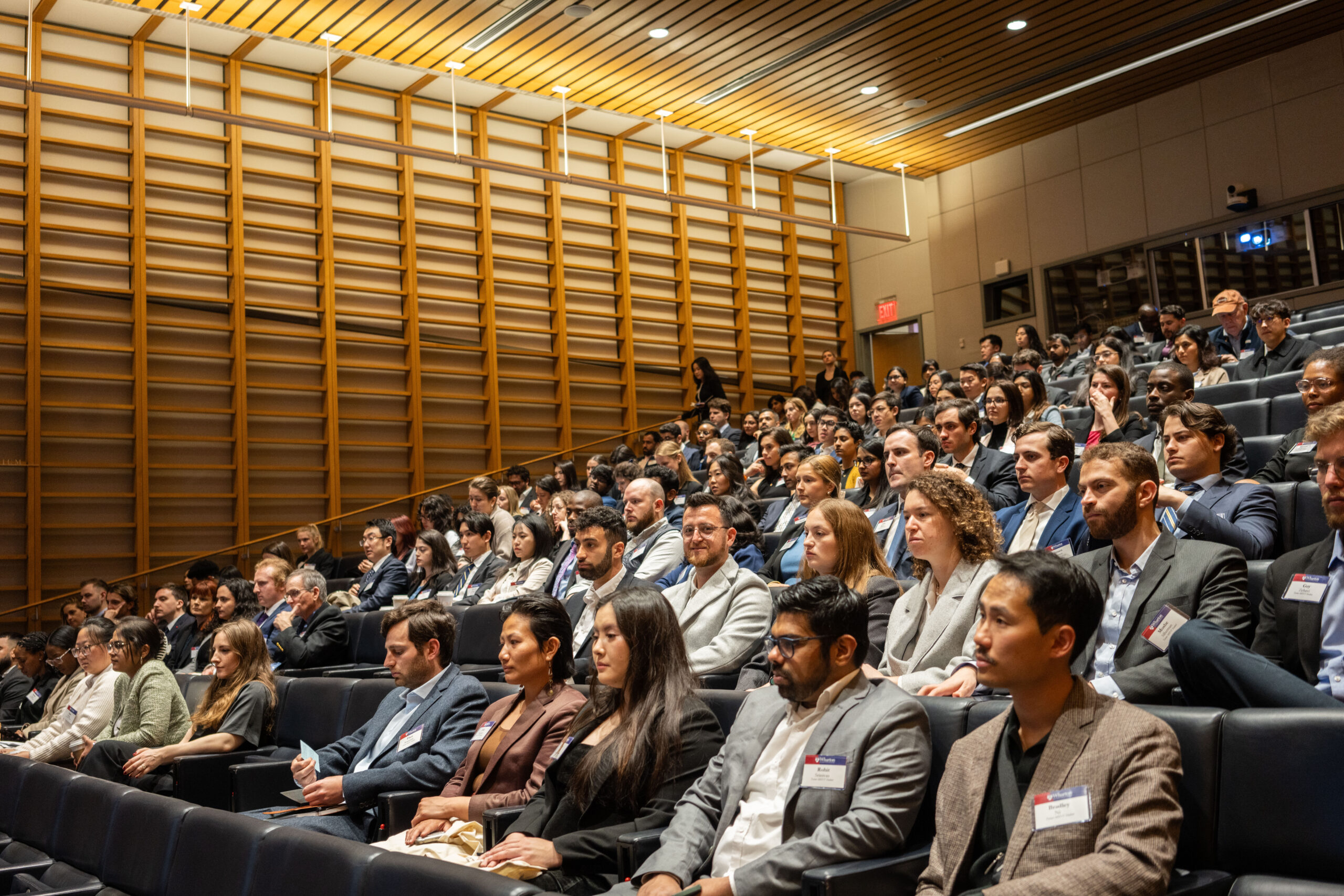 A large group of people seated in a modern auditorium attentively watching something, with wooden paneling and bright overhead lighting.