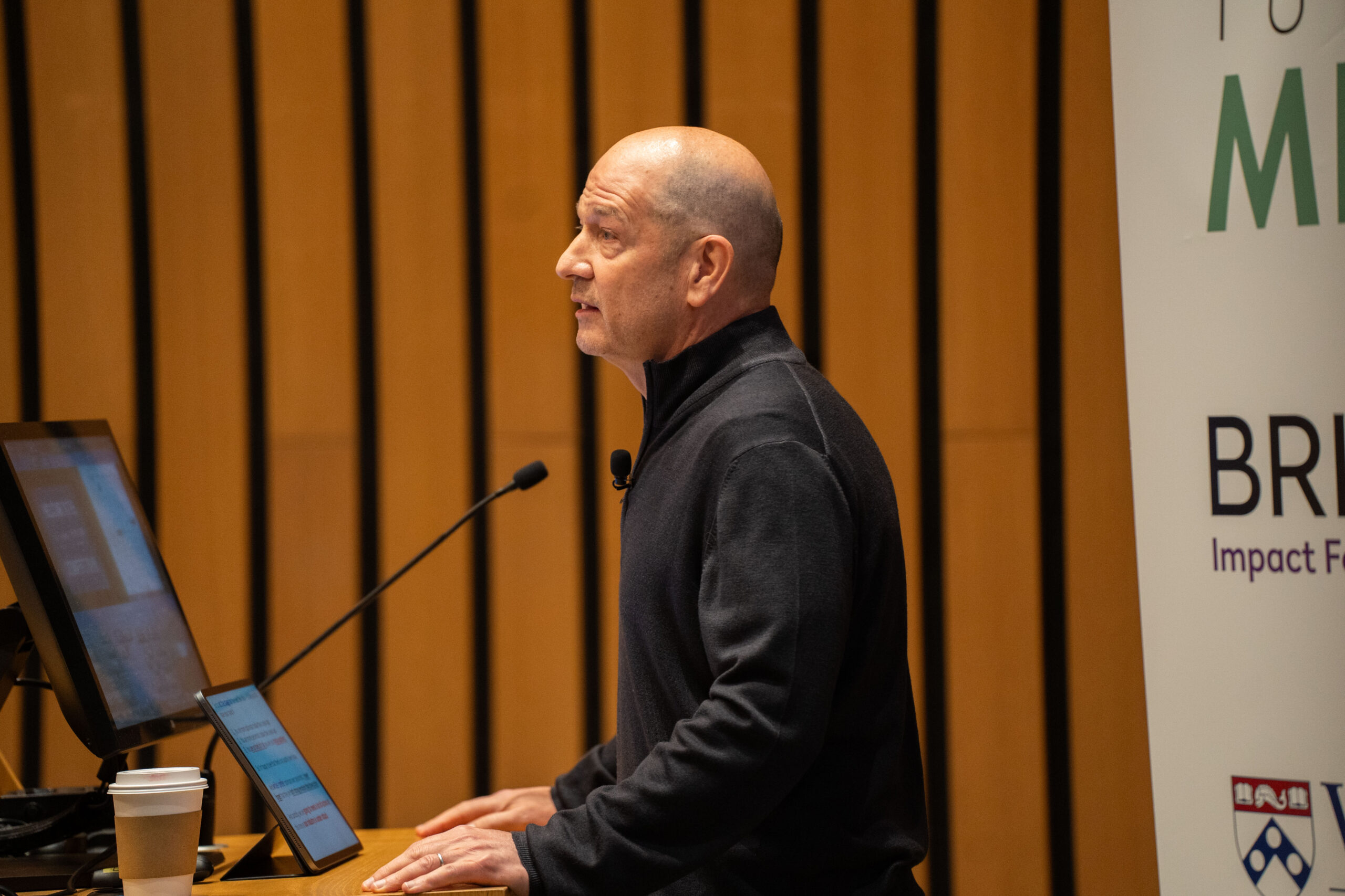 A person is giving a presentation or speech at a lectern, with a computer screen and a microphone visible. There's a coffee cup on the lectern and a banner with partial text in the background.
