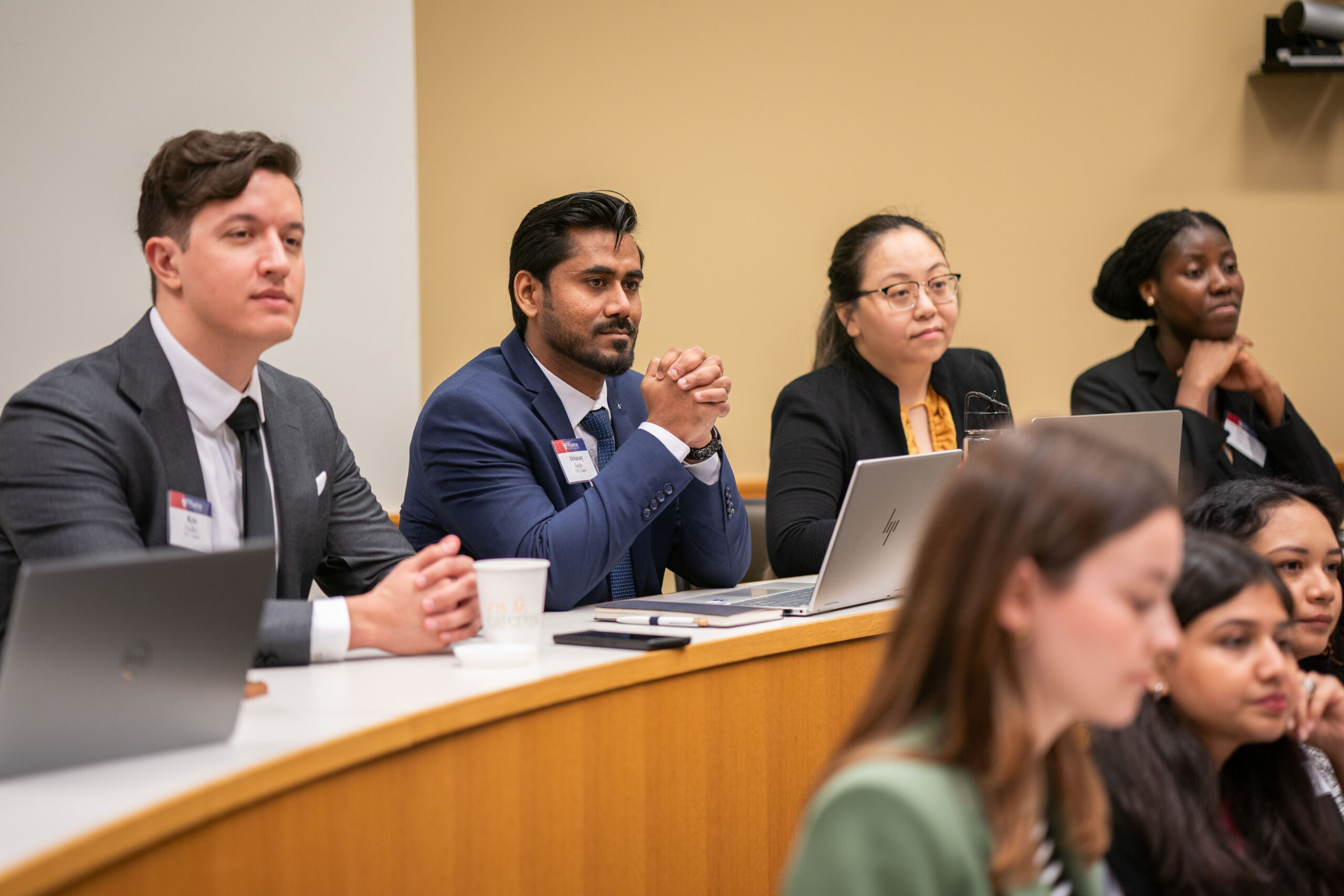 A group of individuals in professional attire are attentively participating in a conference or meeting setting, with laptops and notepads visible.