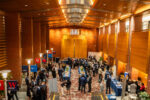 A large group of people is gathered in a spacious, wooden-paneled hall, engaging in a networking event or conference. Multiple tables are set up with banners and promotional materials, suggesting an academic or business setting.