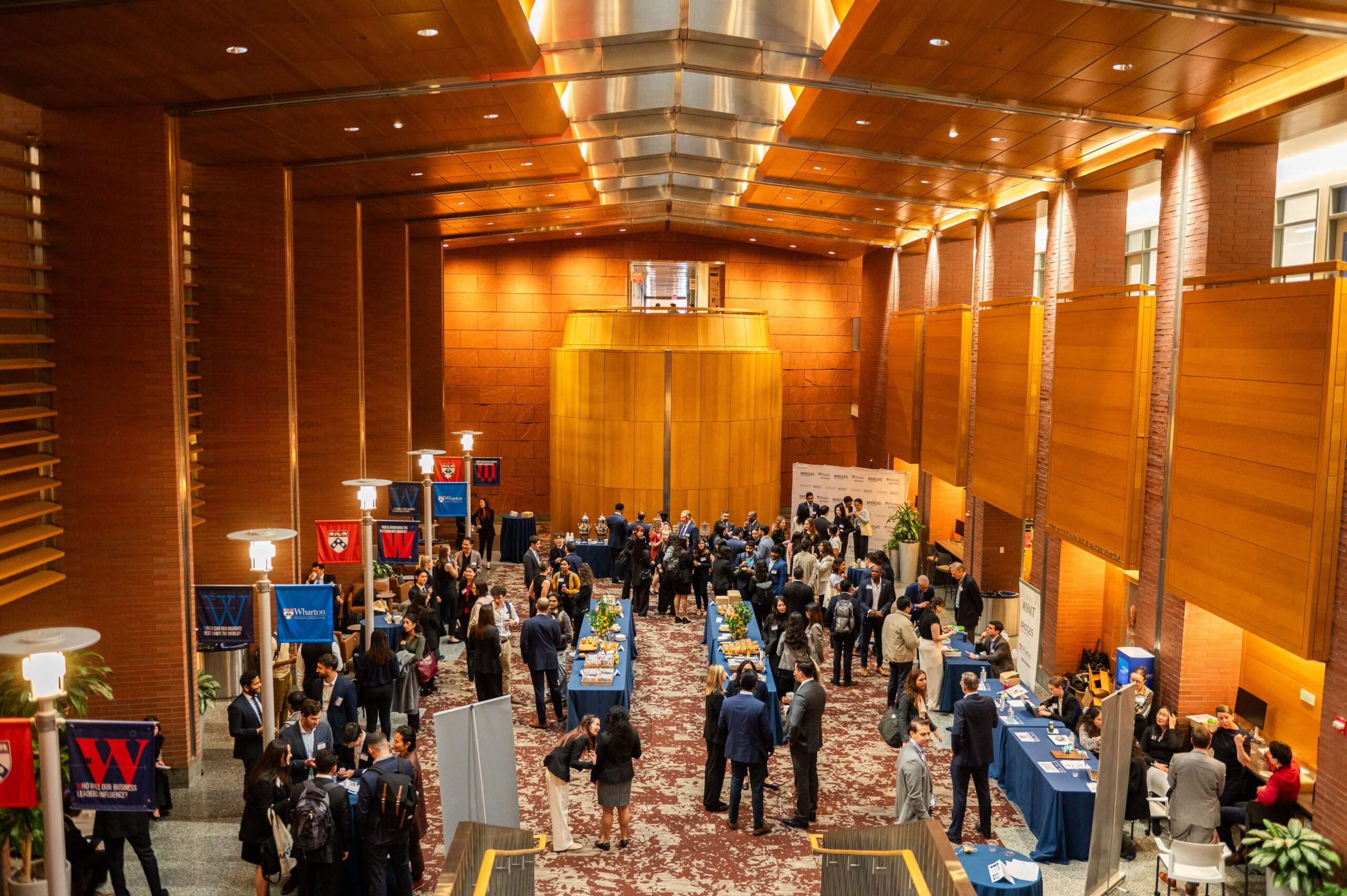 A large group of people is gathered in a spacious, wooden-paneled hall, engaging in a networking event or conference. Multiple tables are set up with banners and promotional materials, suggesting an academic or business setting.