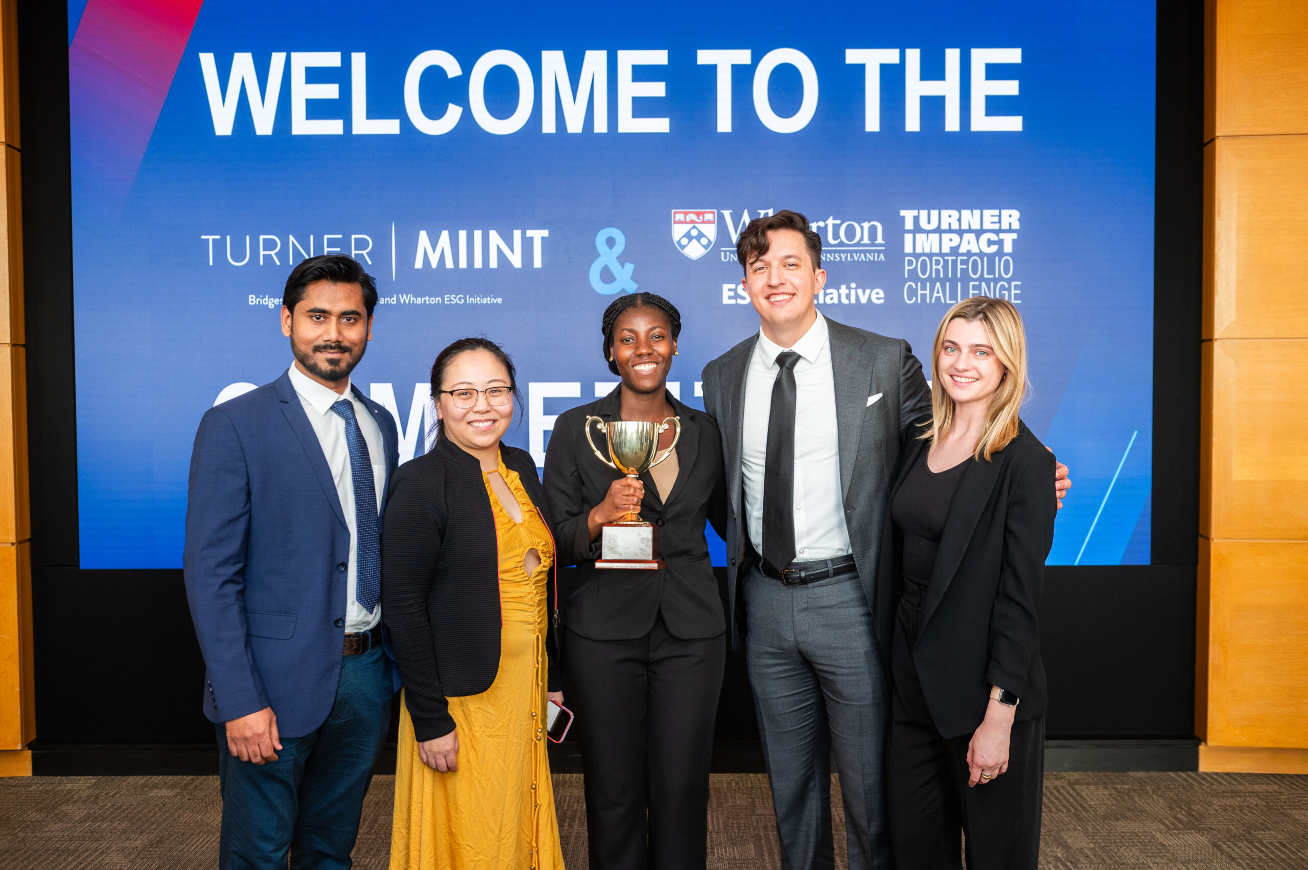 A group of people in formal attire pose in front of a large screen displaying the words "Welcome to the" and various logos. One person holds a trophy, indicating a celebratory or award event.