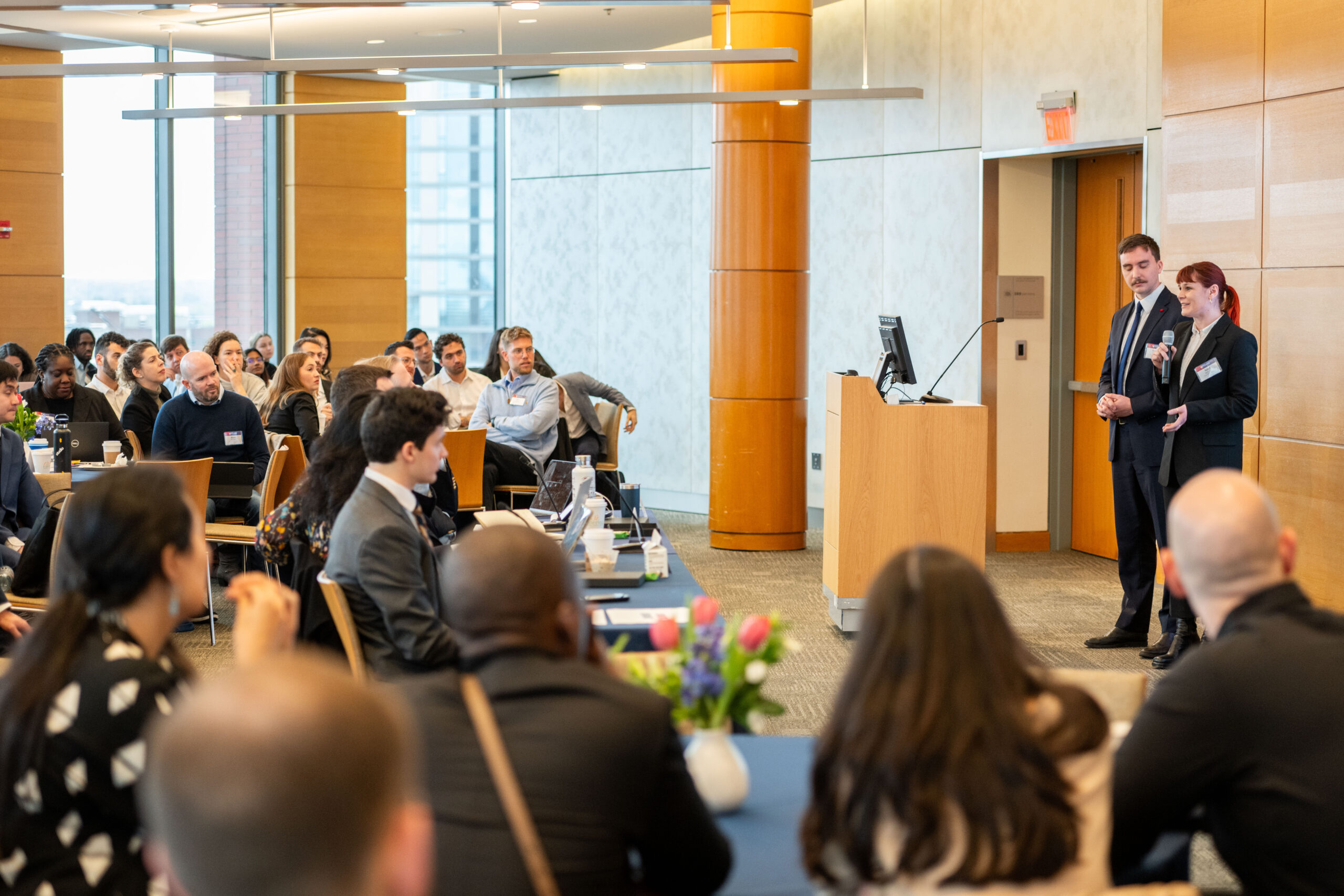 A small group is attending a professional presentation or seminar in a well-lit conference room. Two individuals are speaking at a podium, while the audience listens attentively.