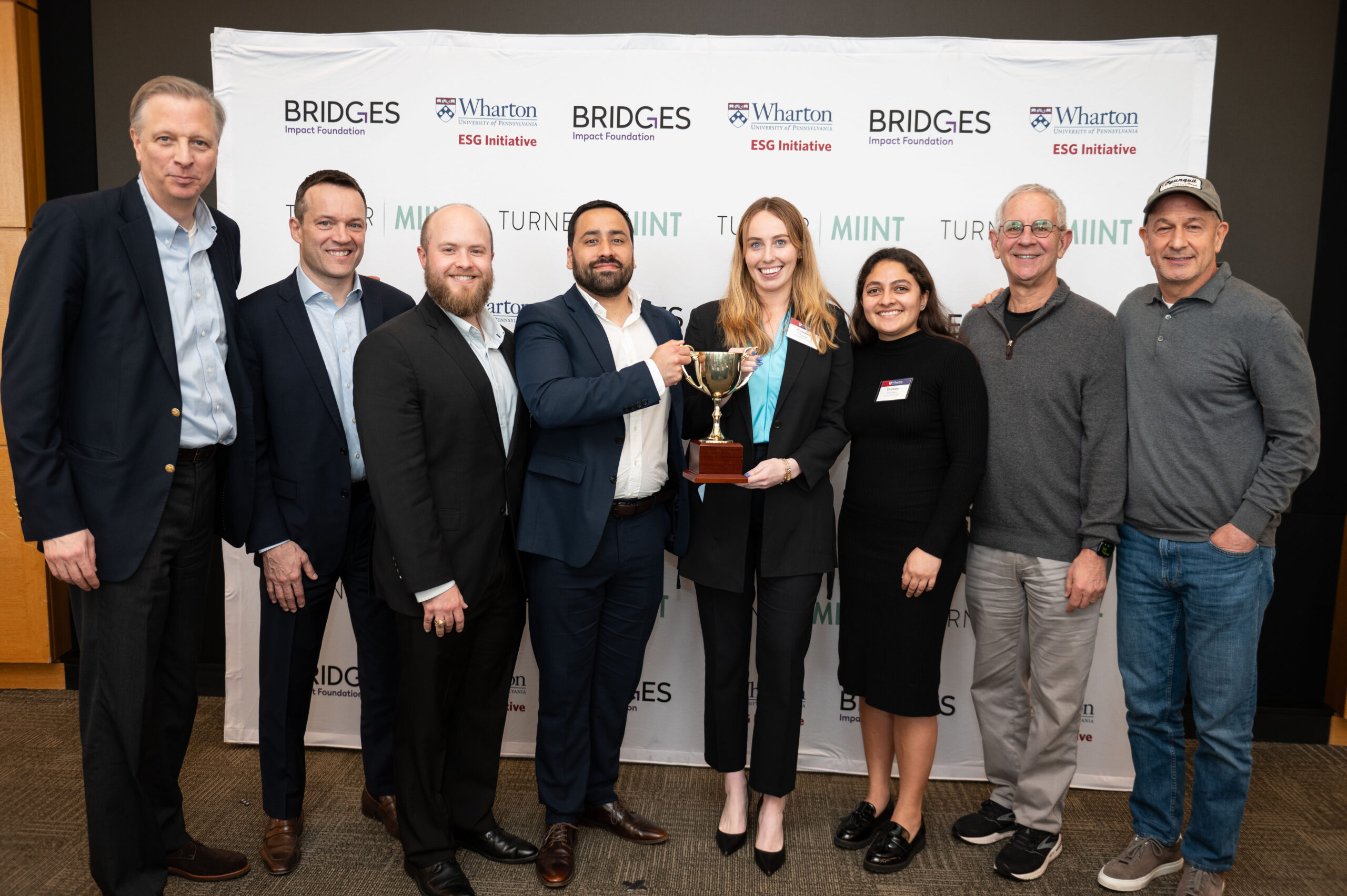 250405 949 A group of eight people in business attire stand in front of a step and repeat with logos, holding a trophy.
