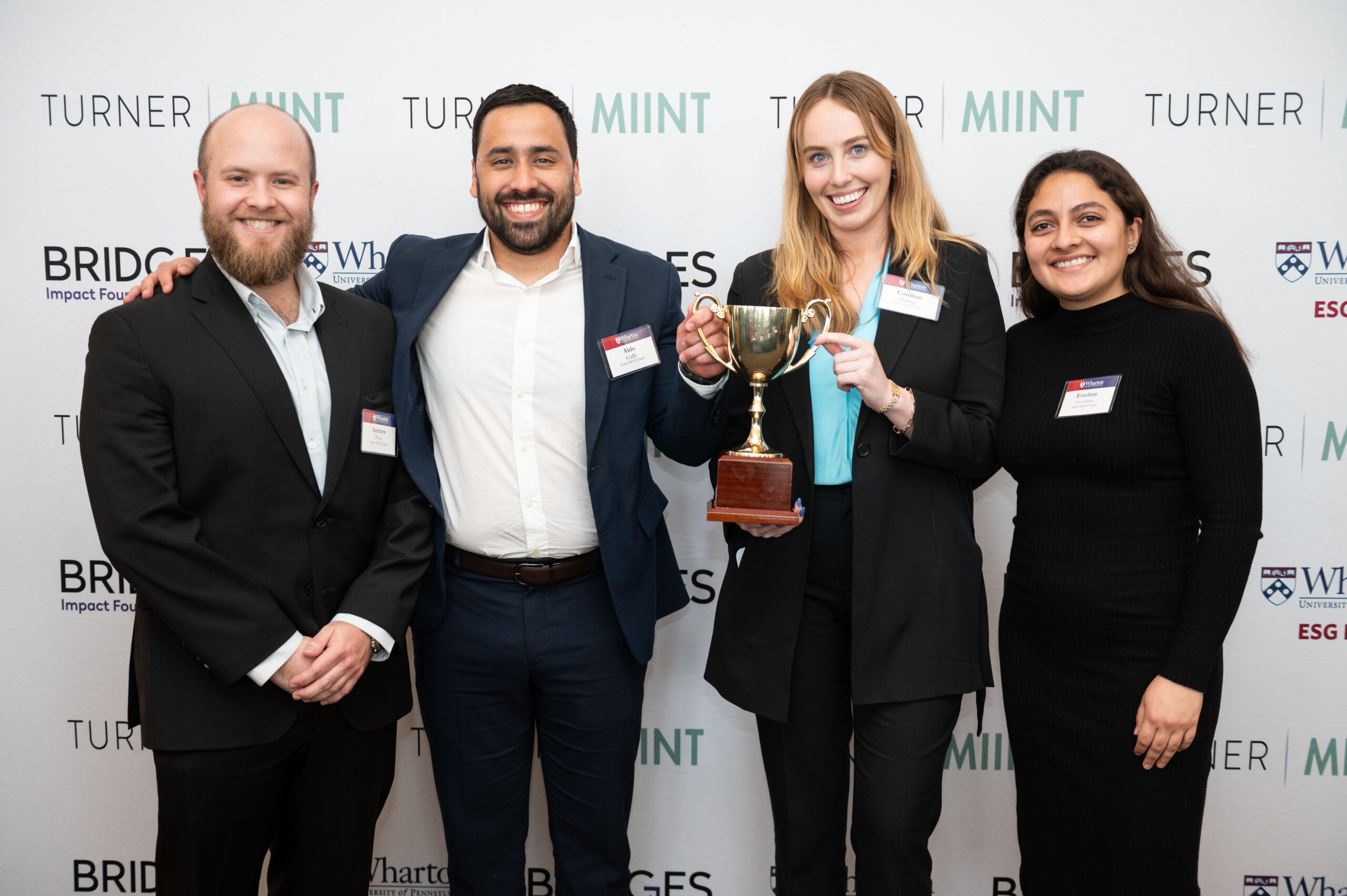 A small group of people in formal attire are smiling at the camera. Two of them are holding a trophy, suggesting a recent achievement or award. A branded backdrop is behind them.