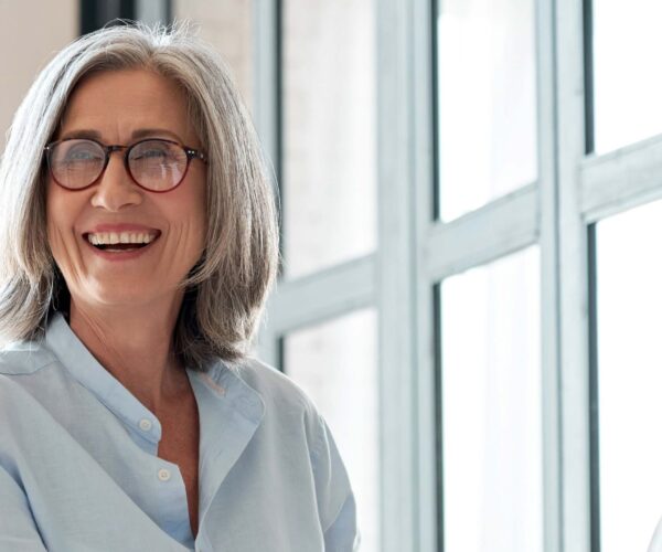 Woman with grey hair wearing a blue collared shirt and red glasses laughs looking off camera in front of a wall of windows.