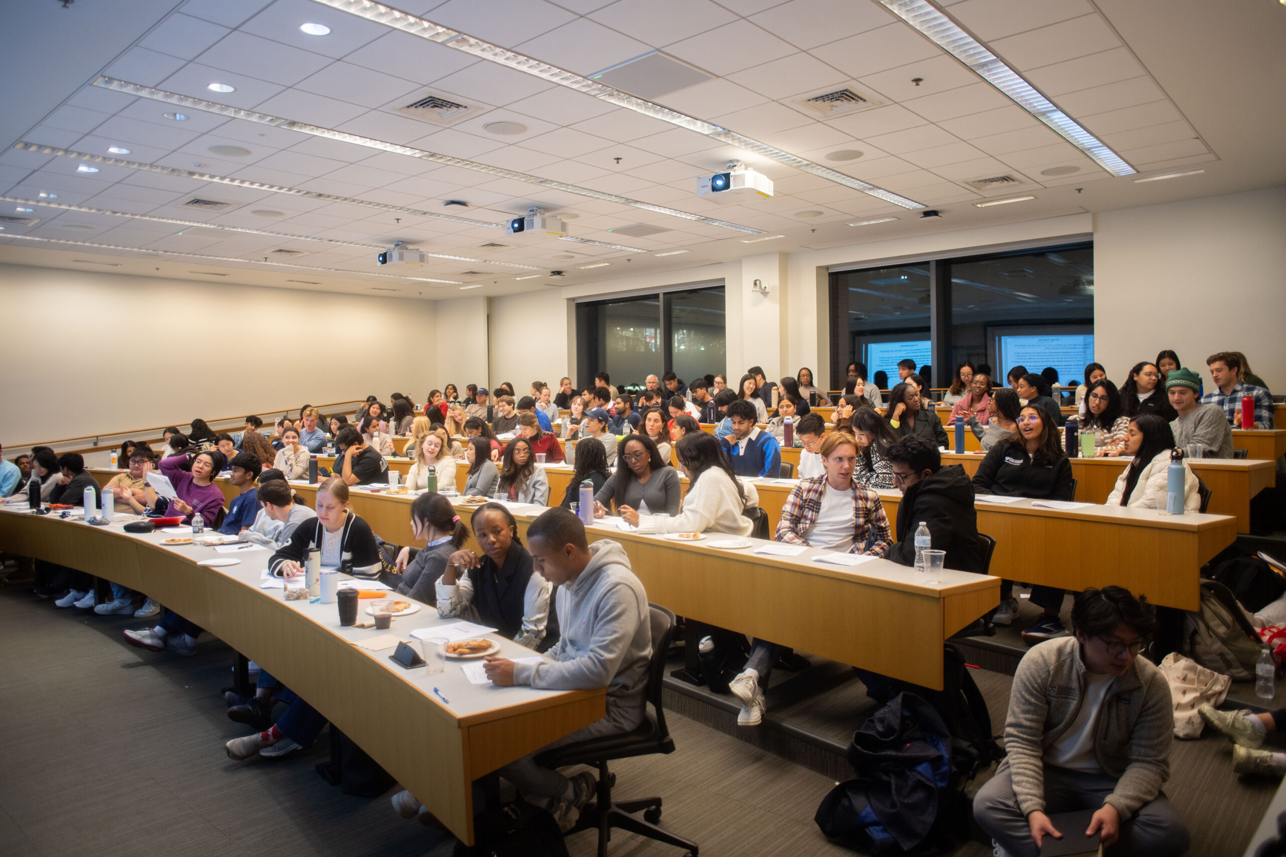 Wide photo of a classroom filled with students sitting at desks and talking